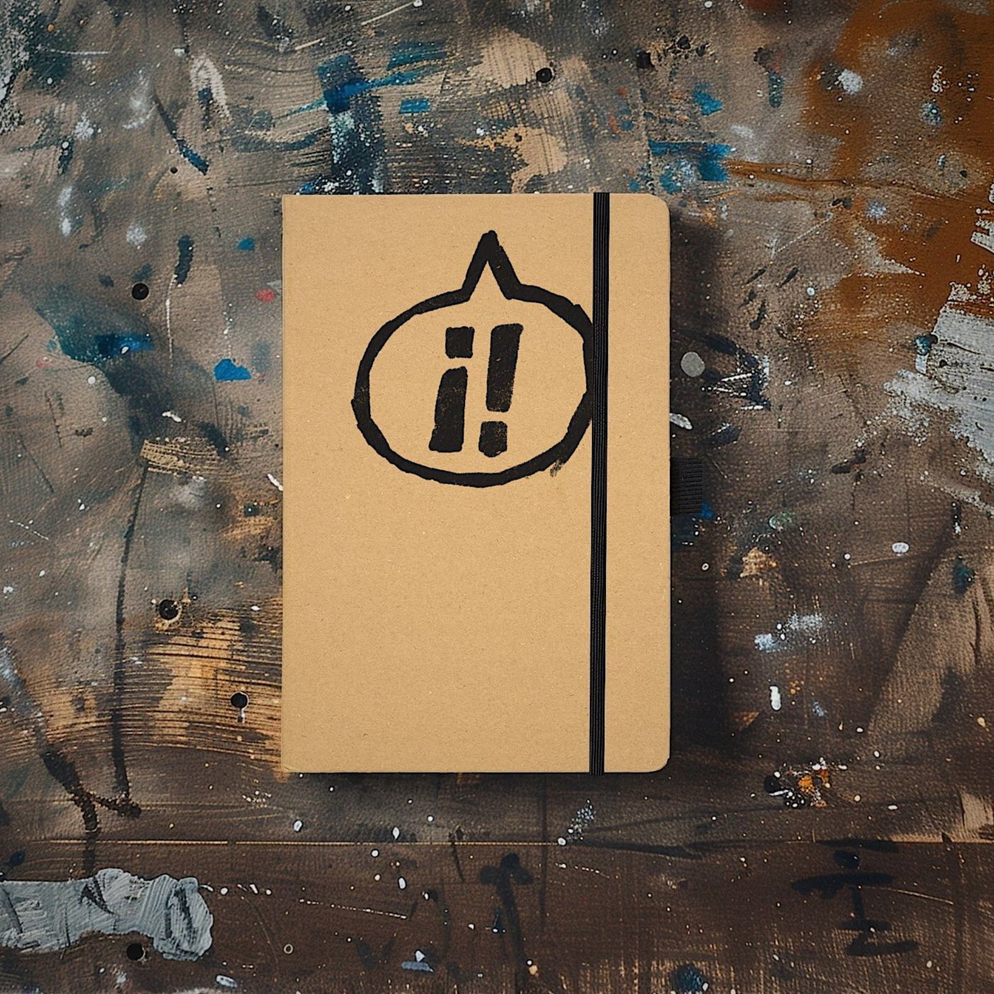 Exclaim Yerself Recycled Kraft paper hardcover notebook, containing 100 pages of lined recycled paper, with recycled closure loop and pen loop, pictured against an artist’s wooden painterly background. The cover is printed with our logo of two exclamation marks ¡! inside a speech bubble pointing to the top of the notebook.