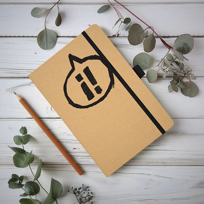 Exclaim Yerself Recycled Kraft paper hardcover notebook, containing 100 pages of lined recycled paper, with recycled closure loop and pen loop, pictured against a white wooden background next to a pencil and some foliage. The cover is printed with our logo of two exclamation marks ¡! inside a speech bubble pointing to the top of the notebook.